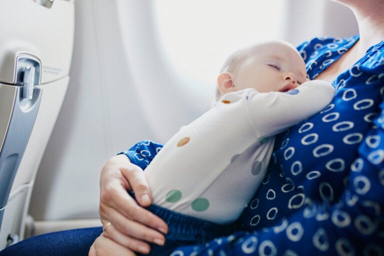 A baby is sleeping in a person's arms, who is sitting by an airplane window. The baby is wearing a white outfit with colorful dots, and the person is wearing a blue shirt with white circles. Traveling with a baby on a plane can be made easier when they're comfortably asleep, as seen here next to the window.