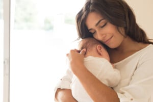 A woman with long dark hair is holding and hugging a newborn baby close to her chest. She is indoors, wearing a white top, and is seen smiling gently while looking down at the baby, who is swaddled in a white blanket. The softly lit background adds warmth to this tender moment as she considers Spanish names for her child.