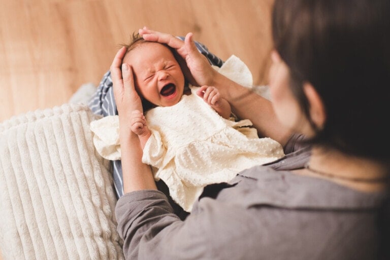 A newborn baby in a white outfit is cradled in an adult's lap. Crying with eyes closed and mouth open, the baby is gently held by the adult's hands supporting its head. Understanding colic can be challenging, but the soothing presence is essential. A white cushion is partially visible beside them.