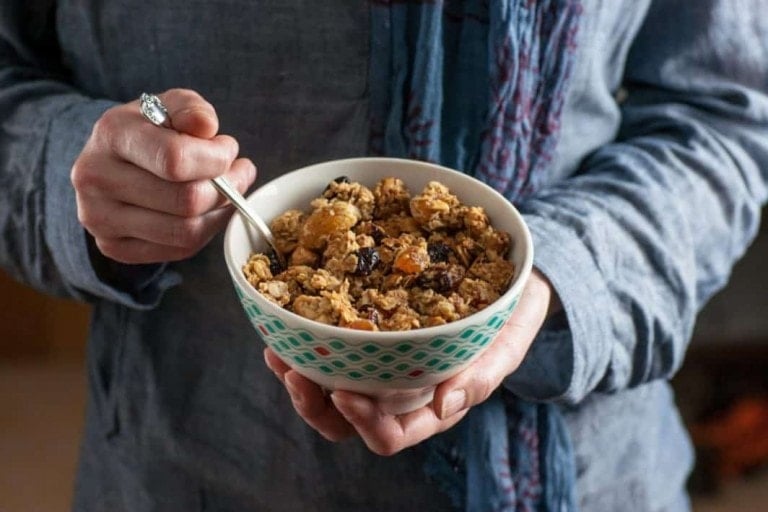 A person holding a bowl of granola with a spoon. The granola, rich in dietary fiber and mixed with dried fruits, possibly raisins, sits invitingly. The person is wearing a blue shirt and a scarf with a ring on their left hand. The white bowl features a green geometric pattern.