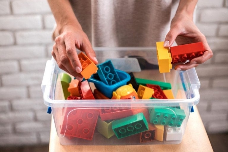 A person is meticulously sorting colorful plastic building blocks in a clear storage container. The container is placed on a wooden surface with a brick wall in the background. The person's hands, aware of the importance of clean kids toys amid COVID-19, are holding orange and red blocks.