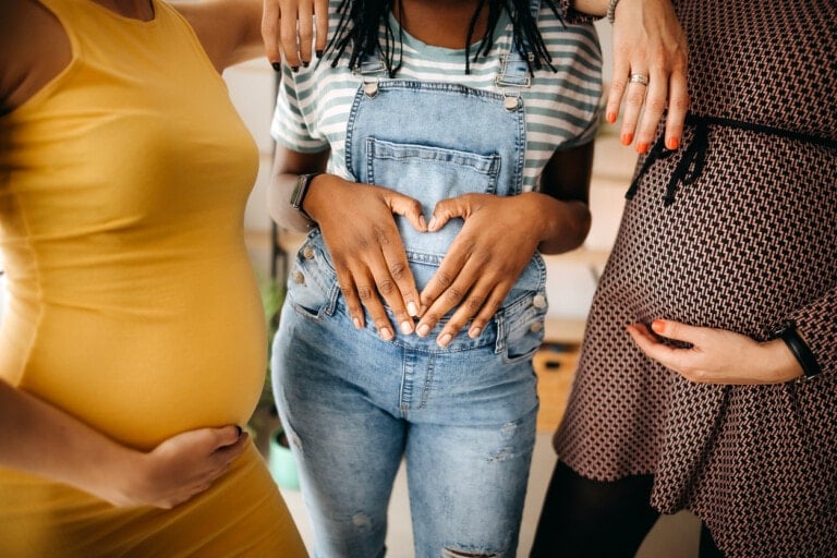 Three pregnant women stand side by side, each with one hand on their belly. The woman in the middle wears denim overalls and a striped shirt, her hands forming a heart shape over her belly. The other two wear dresses—one yellow and one patterned—as they share truths about the second trimester.