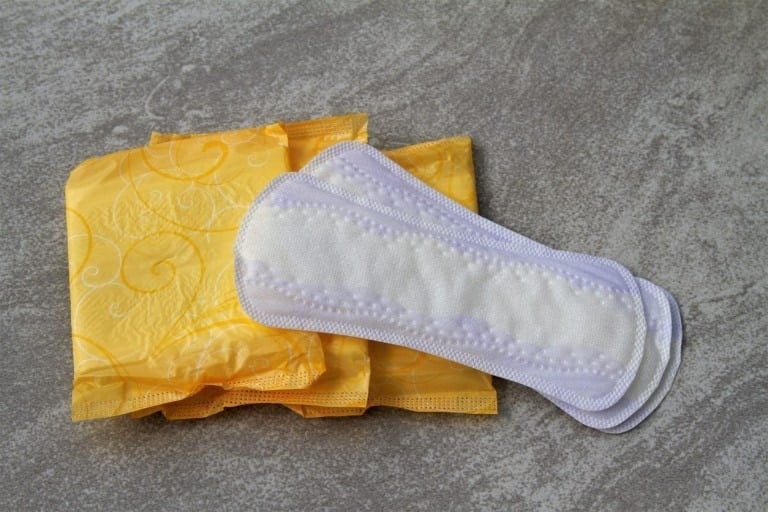 The image shows three sanitary pads: two are wrapped in yellow packaging with a floral design, and one is unwrapped, revealing a white absorbent surface with a light lavender border. They are arranged on a gray textured surface, perfect for creating postpartum padsicles.