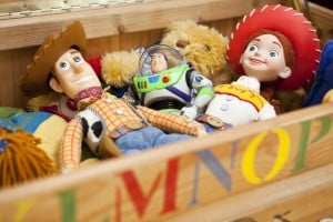 A photo showing three toy characters, including a cowboy, an astronaut, and a cowgirl, lying inside a wooden box filled with various stuffed animals and colorful items. The box has letters painted on its front side, reminiscent of charming Disney baby names.