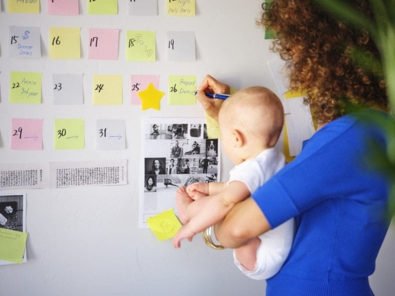 A person with curly hair, wearing a blue top, holds a baby while writing on a sticky note on an organized calendar wall. The wall includes colorful sticky notes, small photos, and handwritten dates. They appear focused on streamlining their tasks.