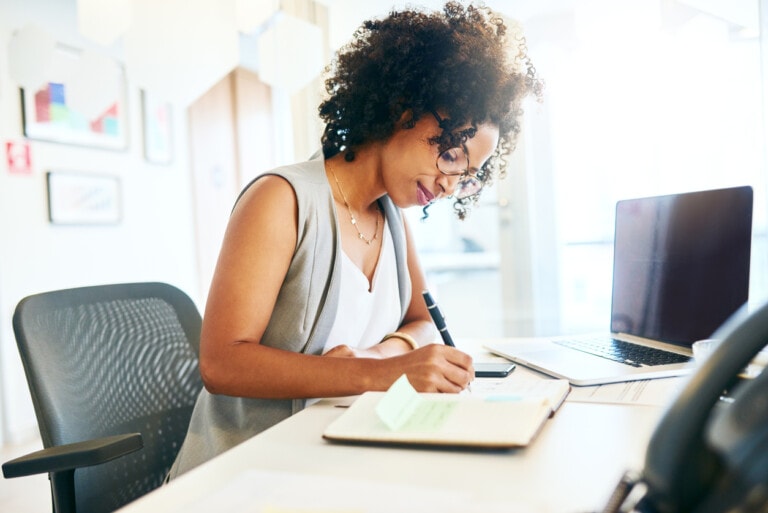 A person with curly hair and glasses is seated at a desk in an office, writing in a notebook. They seem focused on the task, perhaps exploring the benefit of using a planner. There is a laptop, a smartphone, and sticky notes on the desk. The environment is well-lit with light coming from the left side.