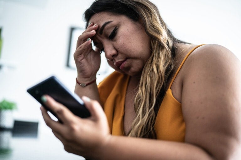 A person with long hair is sitting and holding a smartphone in one hand while resting their forehead on their other, looking concerned. They are wearing a yellow sleeveless top. The background is out of focus, suggesting they might be wanting to call home.