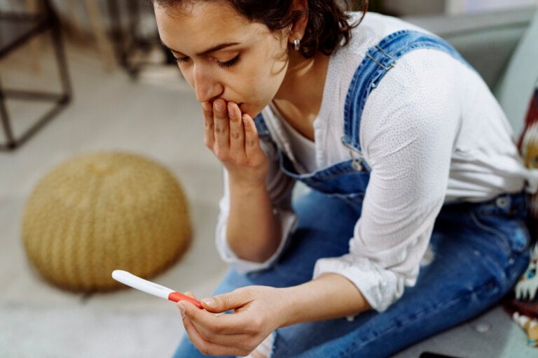 A woman sitting on a couch, wearing blue overalls and a white shirt, holds a pregnancy test in one hand while covering her mouth with the other, looking intently at the result—pondering if it's possible for a stealth pregnancy. A round woven pouf is in the background.