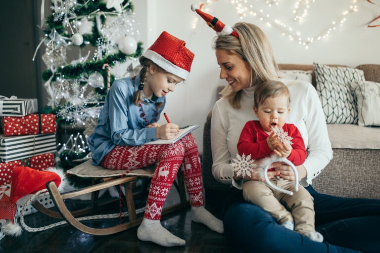 A woman and two children are sitting near a decorated Christmas tree. The older child, wearing festive clothes and a Santa hat, is writing in a notebook while sitting on a sleigh. The woman is smiling at the baby, who is holding a snowflake ornament, capturing the joy of the holiday season.