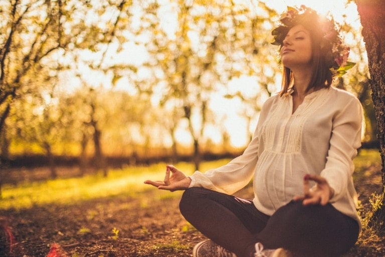 A pregnant woman sits cross-legged outdoors in a sunlit field, meditating with eyes closed. She is wearing a white blouse, black pants, and a flower crown, with sunlight streaming through the trees behind her, peacefully listening to a birth podcast to prepare for labor.