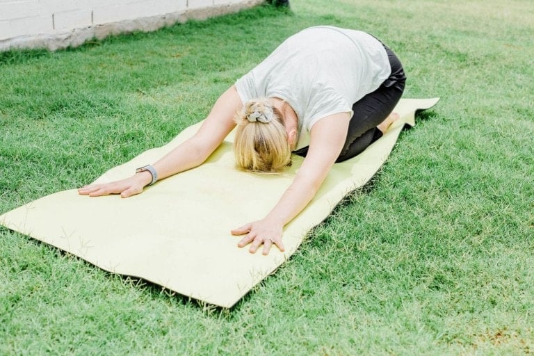 A person with blonde hair, wearing a white shirt and black pants, is performing a postpartum yoga stretch on a yellow mat in a grassy area. They are kneeling with their arms extended forward and their forehead resting on the mat.