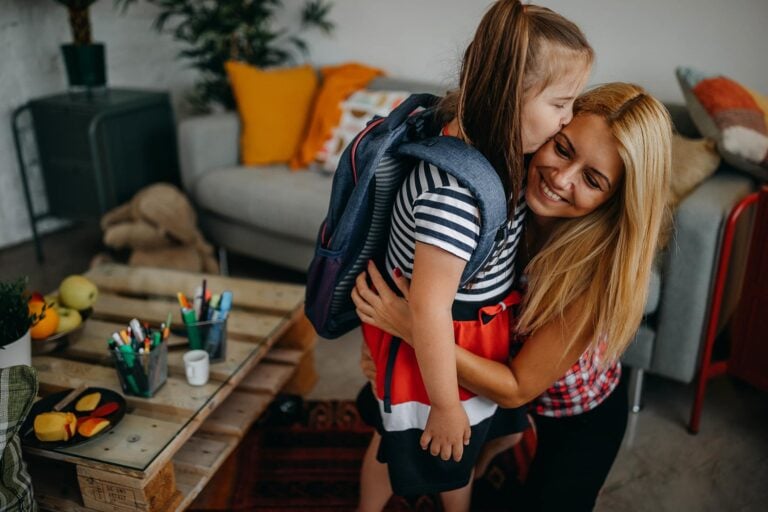 A young girl wearing a backpack hugs a smiling woman with long blonde hair in a cozy living room. The room, designed to help stay organized, features a grey couch with colorful cushions, a wooden coffee table with art supplies, and various decorative items.