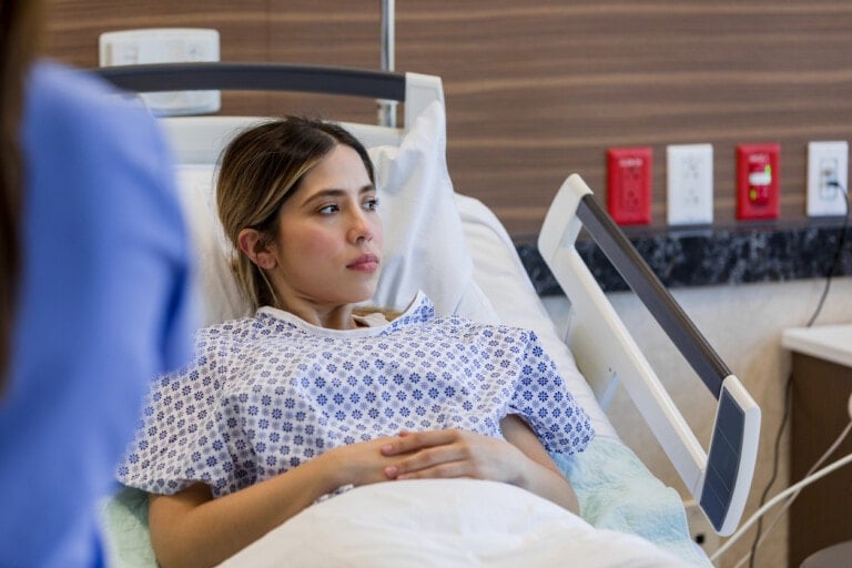 A woman lies in a hospital bed wearing a hospital gown. She appears to be awake and alert, with her hands resting on her stomach. The background includes medical equipment and a wall with various electrical outlets and switches, hinting at the complexity often involved in cases like molar pregnancy.