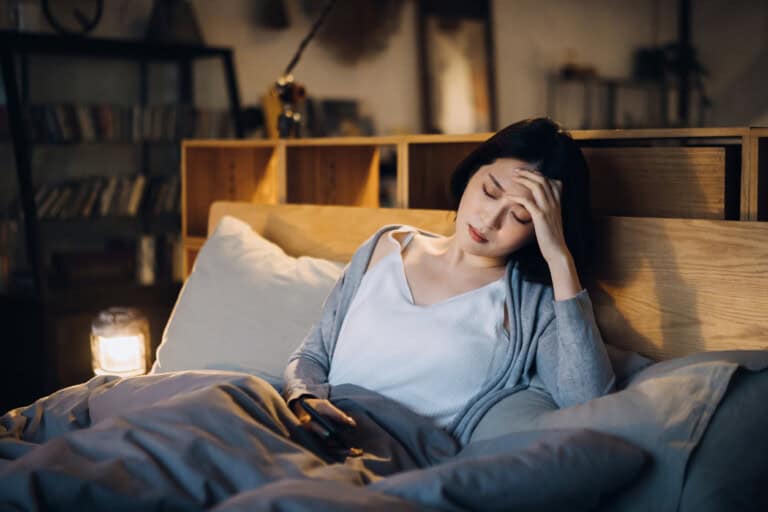 A young Asian woman, looking tired with her hand on her forehead, is sitting up in bed wrapped in a blanket and holding a smartphone. The lit lamp on the bedside table casts a warm glow, while shelves filled with books stand in the background—she shows signs of low iron in pregnancy.