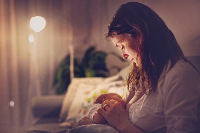 A woman is sitting on a couch in a dimly lit room, breastfeeding a baby, finding support during postpartum. She looks down at the baby with a gentle expression. The room has soft lighting and some plants in the background.