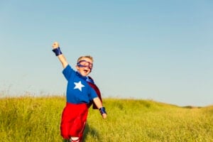 A young child in a superhero costume, perhaps embodying one of those powerful boy names, is running through a grassy field. The child is wearing a blue shirt with a white star, red shorts, a red cape, blue wristbands, and a blue and red mask. The left arm is raised enthusiastically toward the sky.