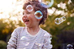 A young child with curly hair and a striped shirt is laughing outdoors, caught in the enchanting chaos of the terrible twos. Several bubbles float in the air around the child, and sunlight filters through trees in the background. The child appears to be enjoying a playful moment.