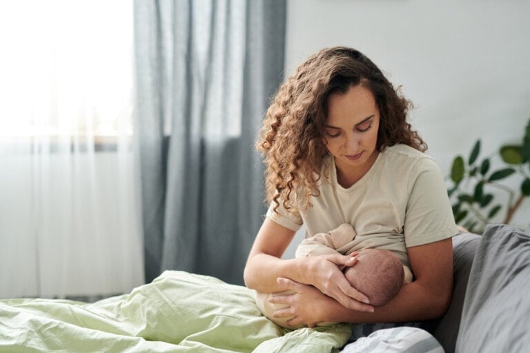 A woman with curly hair, wearing a light-colored shirt, is sitting on a bed and switch nursing an infant. The room has a neutral color scheme, with gray curtains, green bedding, and a plant in the background.