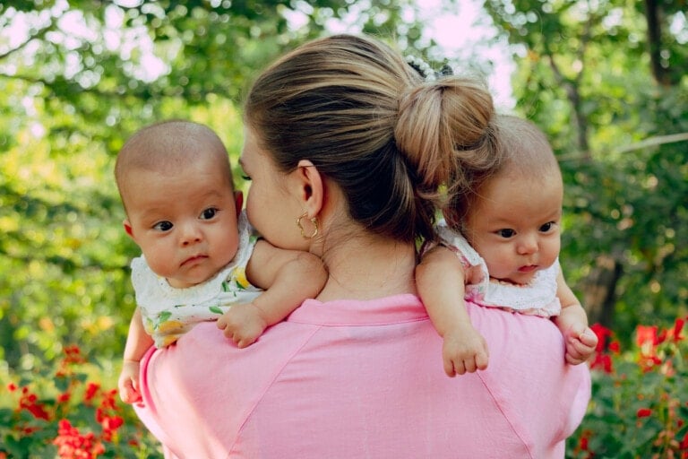 A woman with her hair in a bun wears a pink shirt and holds two babies over her shoulders. The babies, dressed in matching outfits, are looking in different directions—a common occurrence given the intriguing facts about twins. Red flowers and green foliage add to the serene background.