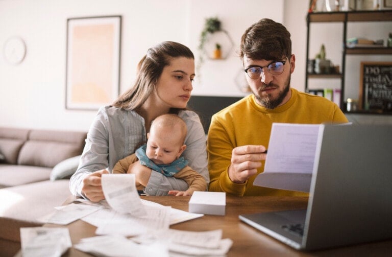 A man and woman, with a baby on the woman's lap, are sitting at a table reviewing documents. The man is holding a paper while the woman examines several in her hand. A laptop and more papers are spread on the table as they focus on financial planning for their child's future.
