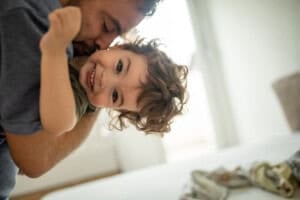 A man is playfully holding a smiling 23-month-old child with curly hair. The child is laughing and facing the camera. The background is softly blurred, showing a bright room and part of a bed, capturing a joyful moment amidst unpredictable sleep schedules.