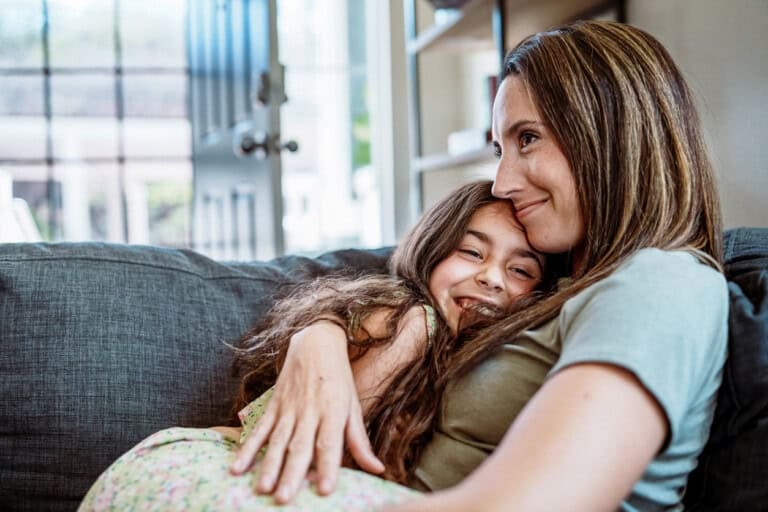 An elementary school age girl sits on her affectionate mother's lap as they cuddle with each other on the living room sofa at home.