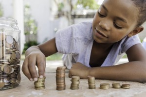 A child is lying on the ground, stacking coins in various piles. A jar filled with more coins sits next to them as they focus on organizing the money. This outdoor setting with greenery and blurred structures provides a perfect backdrop for teaching kids about money.