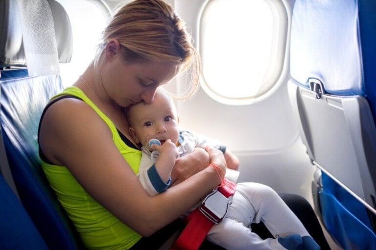 A woman with blonde hair, wearing a bright yellow tank top, is seated next to a window on an airplane. She is holding her lap baby who is sucking on a pacifier. They are both looking down, and the baby is cradled comfortably in her arms.