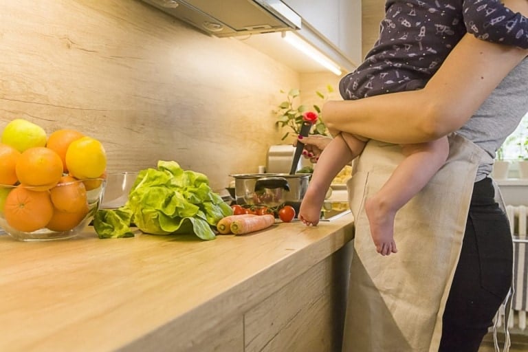 A person holding a small child stands in a kitchen preparing healthy snacks to bounce back after baby. On the counter are various ingredients, including oranges, lettuce, tomatoes, and a carrot. The kitchen has light wooden countertops and cabinetry, with a bowl of oranges placed to the left.