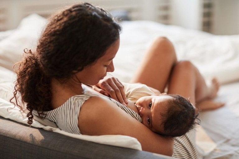 A woman is sitting on a bed, breastfeeding her baby. She is looking down at the infant, who is cradled in her arms. The room is softly lit, creating a warm and peaceful atmosphere—an ideal setting for gifts for breastfeeding moms. Both are dressed in light, comfortable clothing.