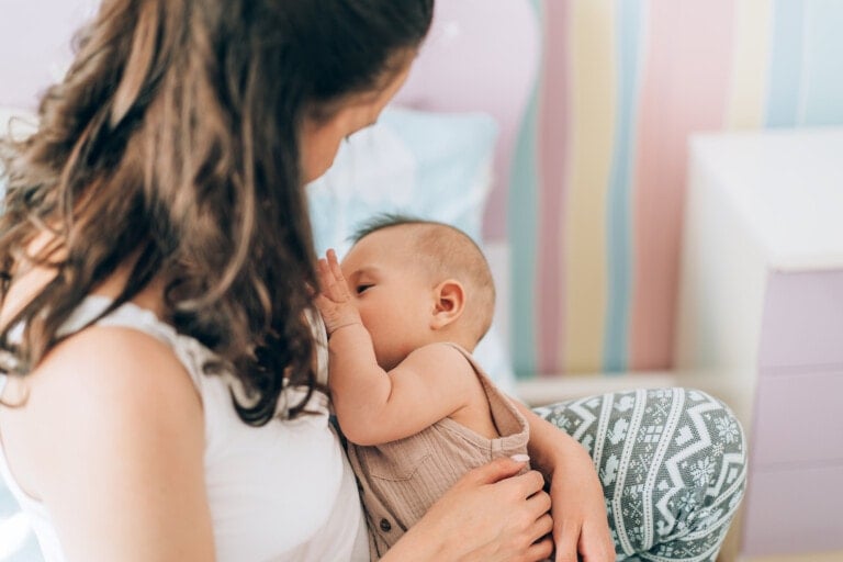 A woman with brown hair is breastfeeding a baby in a room with pastel-colored walls, debunking common myths about breastfeeding. She is wearing a white tank top, and the baby is dressed in a beige onesie. The woman holds the baby close, sitting on a chair or bed.