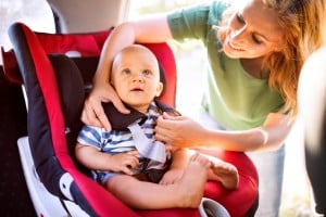A woman secures a fussy baby into a car seat inside a vehicle. The child is seated in a red and black car seat, while the woman adjusts the straps. The baby, wearing a blue and white striped shirt, looks upwards with an expression of mild discomfort.