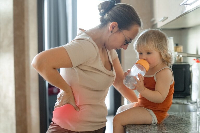A woman stands in a kitchen with her hand on her lower back, delivering one of her parenting pep talks to a young child who is sitting on the countertop, drinking from a sippy cup. The child, with light hair and dressed in an orange outfit, listens attentively.