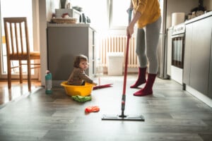 A person mops the floor in a kitchen while a child sits in a plastic tub on the floor. The kitchen has grey cabinets and a wooden chair. Various cleaning supplies, including a bottle of cleaner with potential toxins and cloths, are on the floor nearby.
