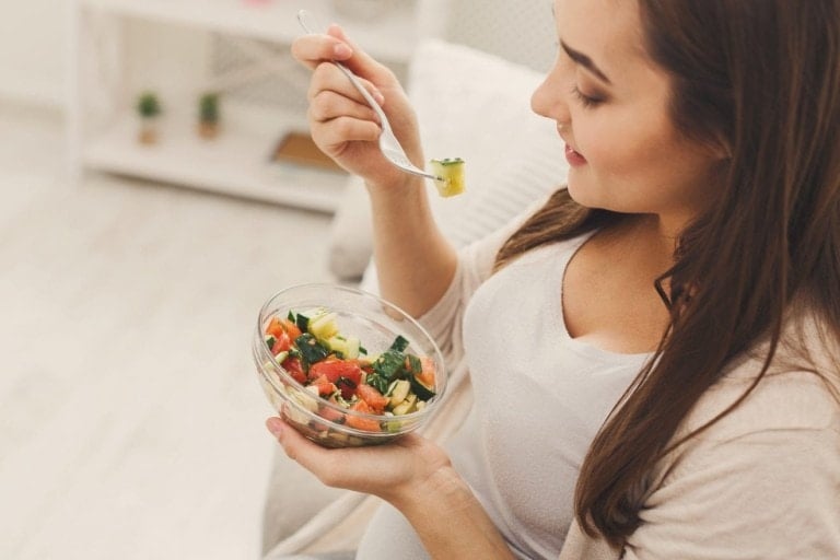 A woman is sitting indoors, holding a fork with food in her right hand and a glass bowl filled with a fresh second trimester recipe salad in her left hand. The salad consists of vegetables such as cucumbers, tomatoes, and leafy greens. She is smiling and looking at the salad.