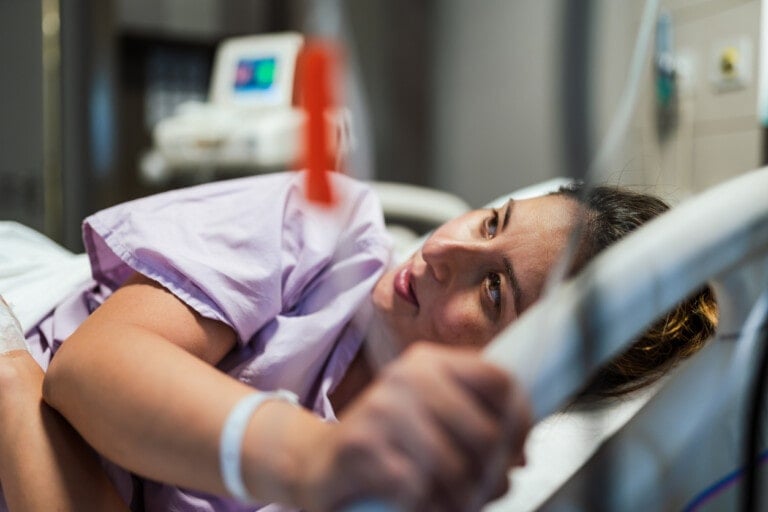 A woman in a hospital bed, wearing a purple hospital gown and a wristband, looks to her left while holding the bed's rail. Connected to an IV line in her arm, she is under observation with medical equipment in the background, possibly preparing for labor induction with Cytotec.