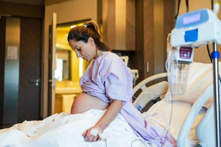 A pregnant person in a hospital gown is sitting on a hospital bed in a patient room. They have an IV catheter in their hand and various medical equipment is visible, including an IV drip. The person appears to be in labor, focusing on their breathing, as they await the next stage of their hospital stay.