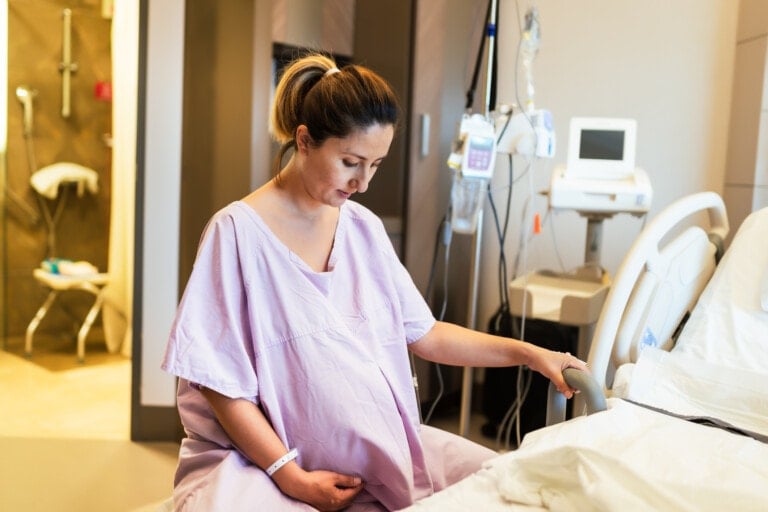 A pregnant woman in a hospital gown sits on the edge of a bed in a labor and delivery room. She holds her belly with one hand and rests her other hand on the bed railing. Medical equipment and monitors are visible in the background.