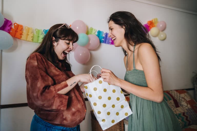 In a room filled with colorful balloons and banners, two women share a joyful moment. One, who is pregnant wearing a brown top, eagerly receives a gift bag with gold polka dots from her friend in a green dress. Its clear that baby shower gifts make the occasion even more special for these happy friends.