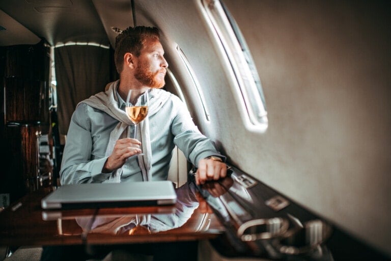 A man with a trimmed beard sits in a private jet, wearing a light blue shirt with a grey sweater draped over his shoulders. He holds a glass of white wine and gazes out the window, seemingly unconcerned that dad leaves kids in coach. A closed laptop is on the table in front of him.