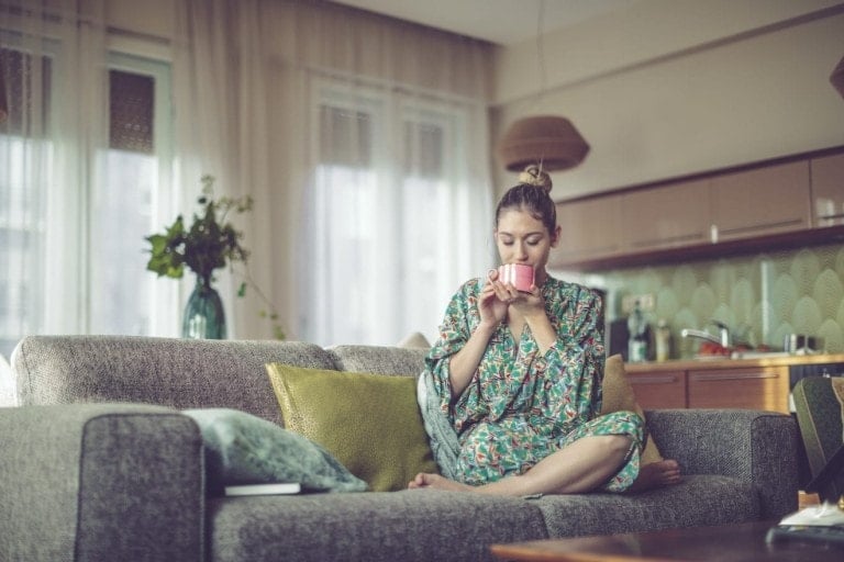 A woman sits on a grey couch in a modern living room, wearing a green floral robe and sipping from a pink mug. The room, ideal for self-care for moms, features large windows, plants on a table, and a kitchen area with light brown cabinets and a patterned backsplash in the background.