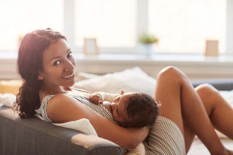 A woman is reclining on a couch and breastfeeding an infant, showcasing the benefits of breastfeeding. She is wearing a striped dress and smiling at the camera. The setting appears to be a well-lit living room with large windows in the background.
