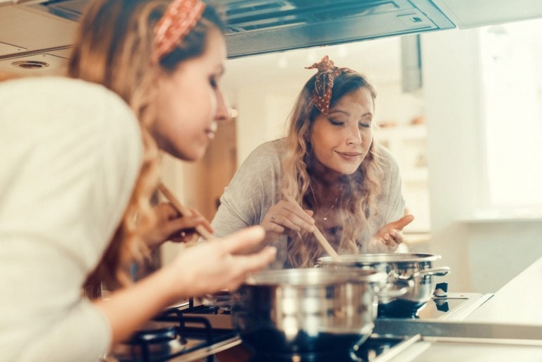 A woman with a headband is cooking in a kitchen, carefully stirring a family-friendly soup recipe in a pot on the stove. She is gazing into the pot and appears to be smelling the food. Her reflection is visible in the background, showing a sense of concentration and enjoyment.