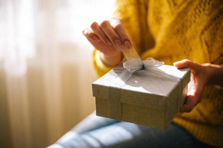 A person wearing a yellow sweater is opening a gray gift box with a white ribbon, their hands slightly trembling. The image focuses on their hands and the gift box, with light from the window casting soft shadows in the background. This moment carries an extra weight, as they are struggling with infertility.