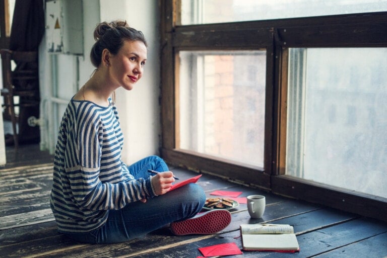 A woman sits on a wooden floor by a large window, writing in a red notebook about what to write in a baby shower card. She is dressed in a striped sweater and jeans, with her hair in a bun. Next to her are a cup of coffee, a plate of cookies, and an open book titled "Baby Shower Card Ideas.