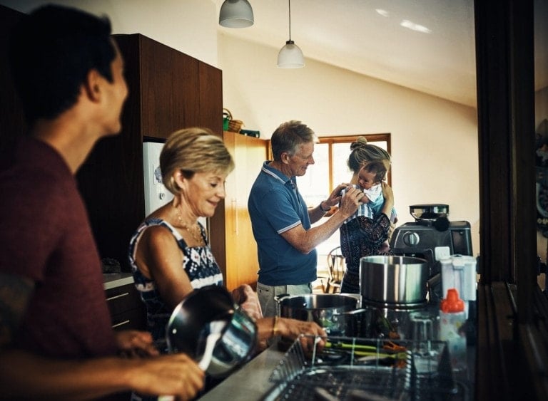 A group of four people in a kitchen. An older man holds a child and plays with them while a woman watches, showcasing how to help when visiting a new mom. Another man is holding a pot, smiling. Various kitchen appliances and utensils are on the counter. The setting appears to be bright and modern.