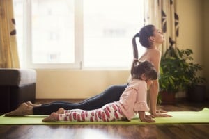 An adult and a child are doing yoga on a green mat inside a room with wooden floors and large windows. Both are in a cobra pose, one of the popular kids yoga poses. The adult's hair is tied back, and the child is wearing a pink shirt and leggings with a watermelon pattern.