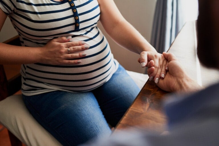 A pregnant woman sits holding her belly with one hand and another person's hand with the other. She is wearing a striped shirt and jeans, while the person whose questions to ask before having a baby she holds hands with is partially visible, wearing a blue shirt.