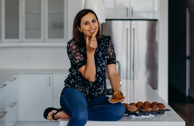 A woman sits on a kitchen countertop eating a muffin, embodying the relaxed, homey vibes championed by the Feel Good Foodie. Casually dressed in jeans and a patterned blouse, she enjoys her treat with a tray of muffins in front of her, and kitchen cabinets and a refrigerator visible in the background.