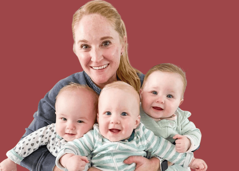 A mom of triplets with red hair is smiling while holding her three babies. Two are dressed in light green, and one wears a polka-dotted outfit. The background features a solid maroon color.